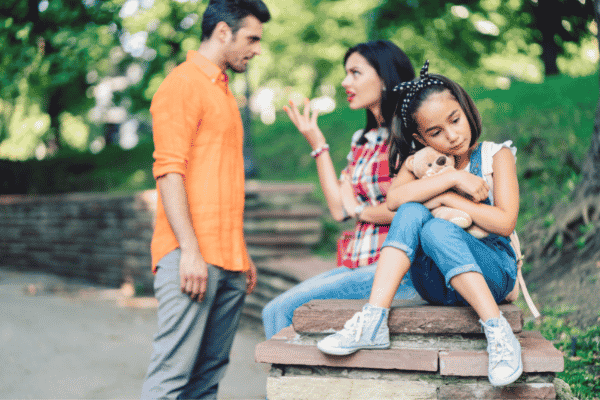 Young girl sitting on a brick wall looking sad while she holds her teddy bear and her parents are arguing behind her
