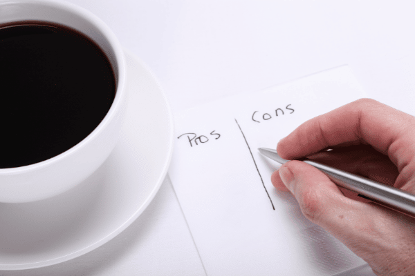 photo of a white coffee cup full of black coffee with a hand writing a pros and cons list on a napkin