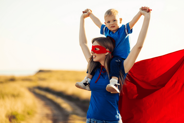 Mom dressed in a blue t-shirt with a red superhero mask and red superhero cape has her son sitting on her shoulders holding both his hands up and out towards the sky
