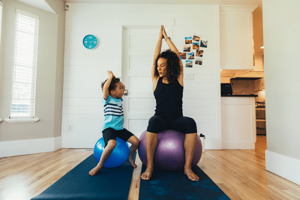 Mother and son each on a yoga ball with their feet on the floor and their hands above their heads with palms facing inward practicing balance
