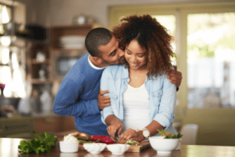 African American husband and wife in the kitchen together, wife is chopping food and smiling and husband is kissing his wife on the cheek